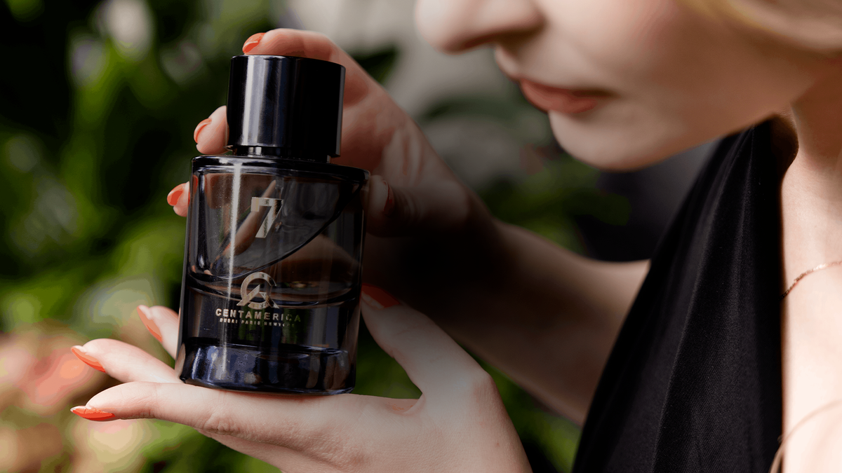 A woman in a black top holds a dark glass perfume bottle with a black cap. The bottle has gold lettering that reads "7 by Centamerica".